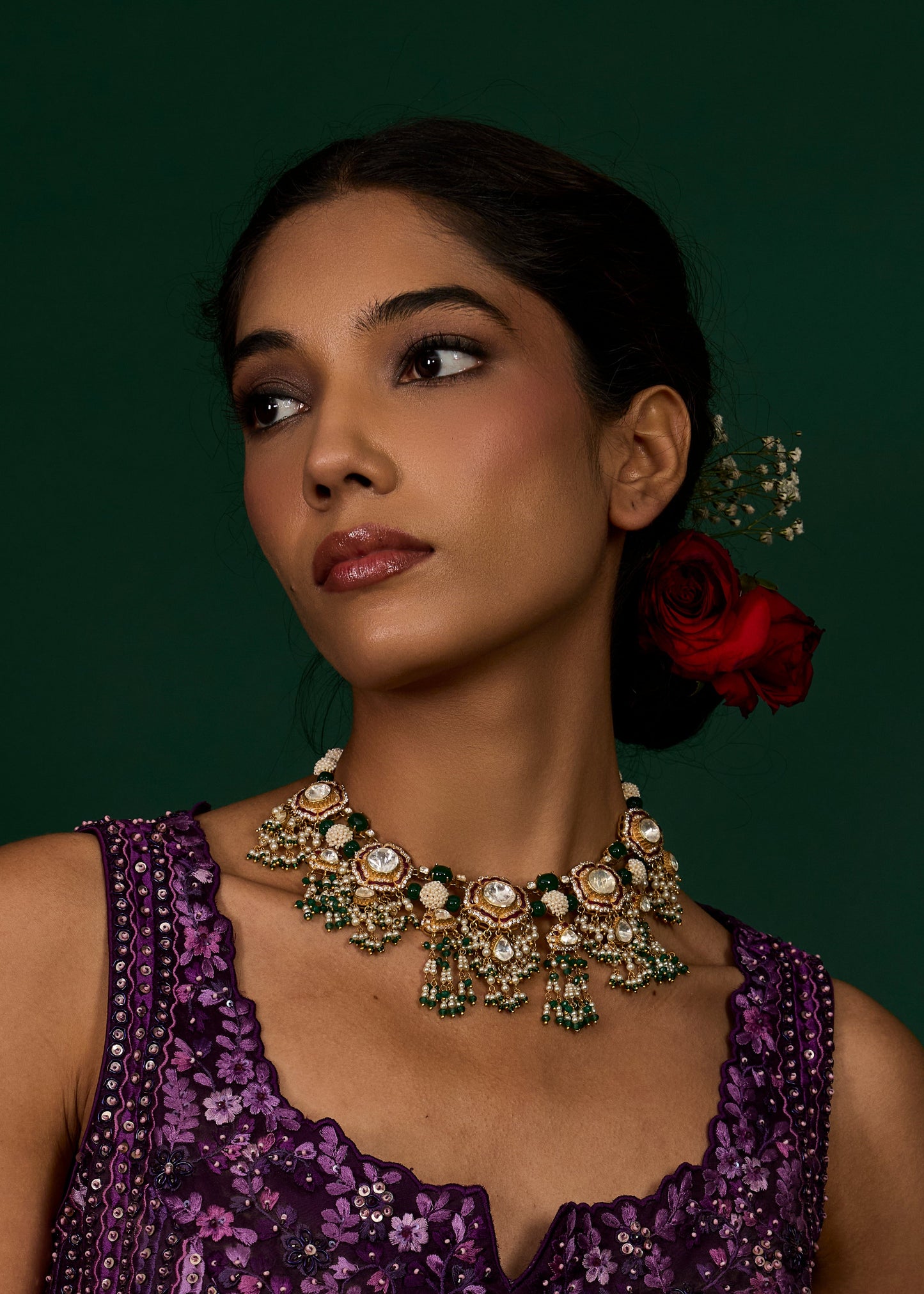 Woman wearing a purple floral dress and ornate jewelry with a red flower in her hair against a dark green background